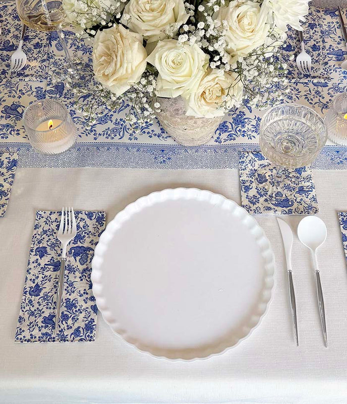 A Royal Peacock Table Setting: Eco White & Blue by Sophistiplate features an elegant white plate, floral napkins, silverware, glassware, a candle, and white roses with baby's breath on a blue and white runner.