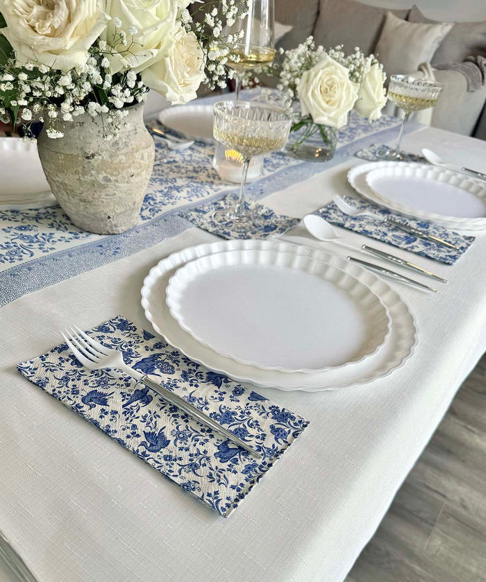 The Royal Peacock Table Setting: Eco White & Blue by Sophistiplate features designer disposable plates, blue and white floral napkins, silver cutlery, crystal glasses, and a centerpiece of white roses in a stone vase for an elegant party-in-a-box look.