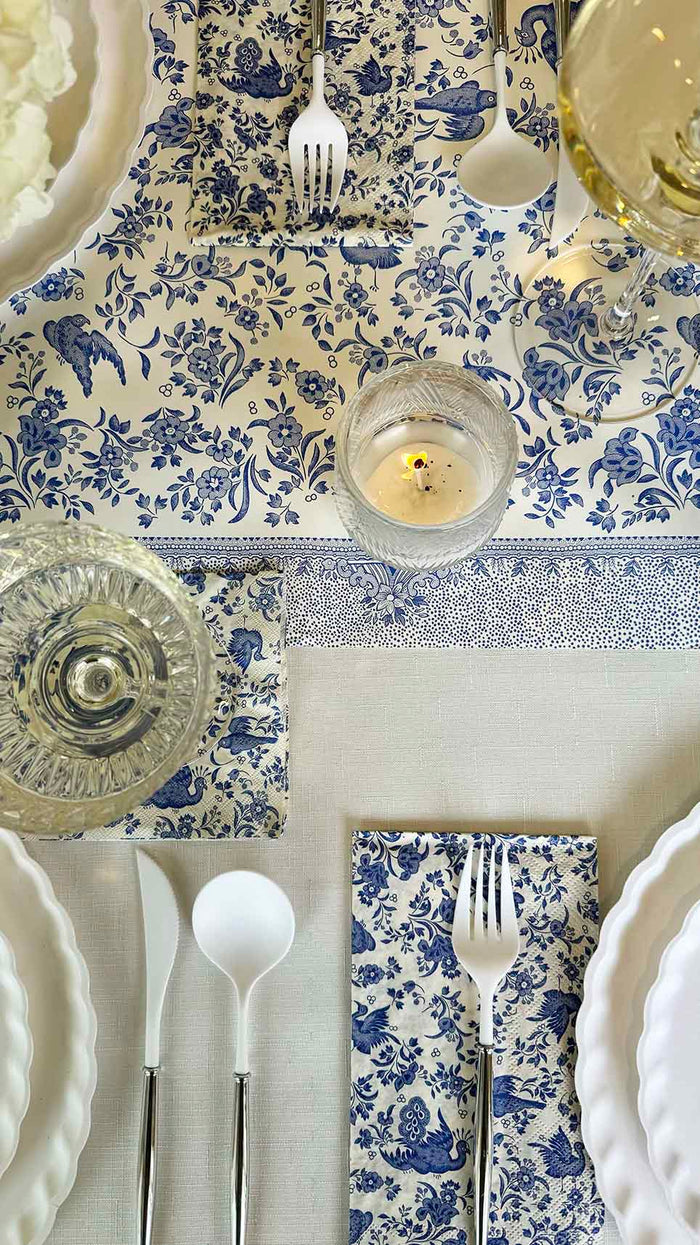 The Royal Peacock Table Setting: Eco White & Blue by Sophistiplate features designer disposable plates, silverware, and wine glasses on a white and blue floral tablecloth and napkins, centered with a candle and glass of water.