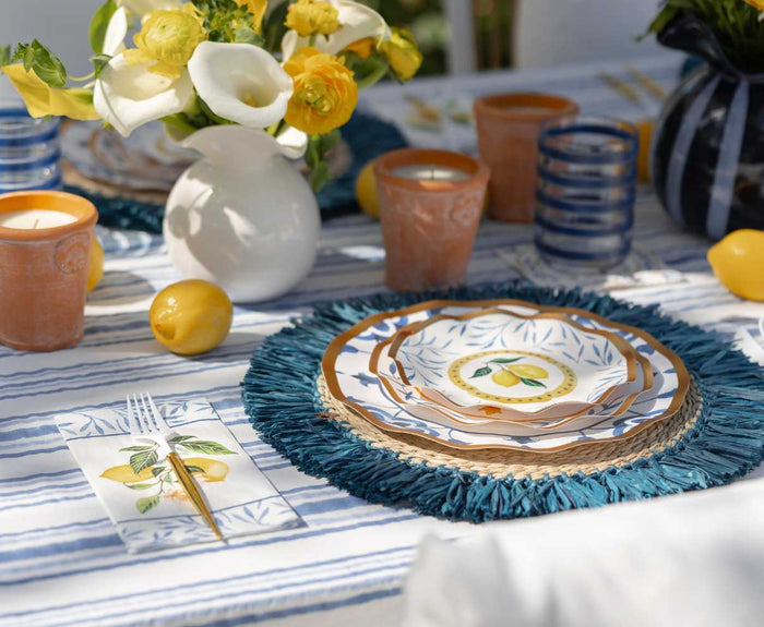 Create a vibrant summer look with the Capri Coast Table Setting by Sophistiplate LLC, featuring lemon-themed plates, a blue fringed placemat, glassware, terracotta candle holders, and a white vase of yellow blooms on a striped tablecloth with scattered lemons.
