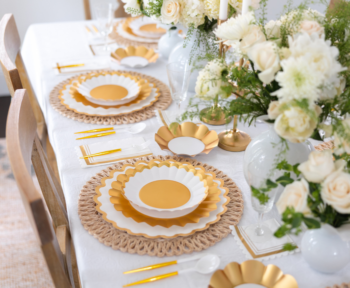 The Gold & White Table Setting by Sophistiplate LLC displays designer disposable plates with gold and white scalloped edges, gold utensils, white napkins, and floral centerpieces on a classic white tablecloth.