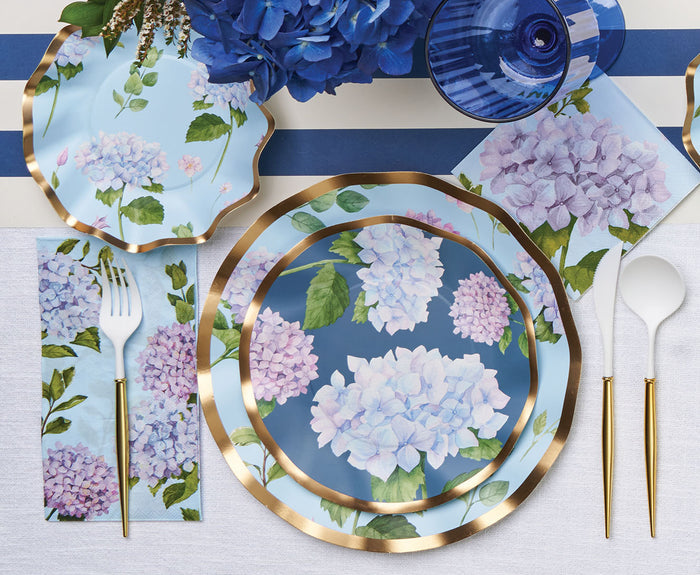 The Hydrangea Table Setting by Sophistiplate features designer disposable plates and napkins with pastel blue, purple, and white hydrangea designs with gold edges, plus a fork and spoon—perfect for an elegant floral-themed table.