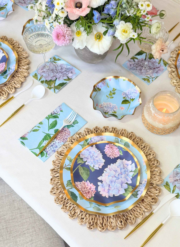 The Hydrangea Table Setting by Sophistiplate features designer disposable plates, gold flatware, a candle, and fresh assorted flowers on a light tablecloth—creating an inviting, elegant spring atmosphere for any occasion.