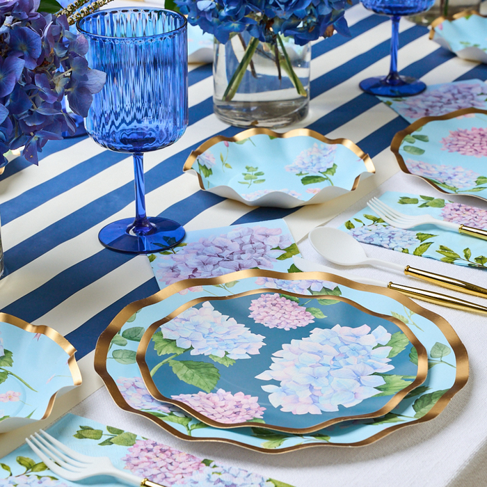 The Hydrangea Table Setting by Sophistiplate features a blue and white striped tablecloth, blue glassware, hydrangea vases, and designer disposable plates that beautifully mimic gold-rimmed floral china for stylish gatherings.