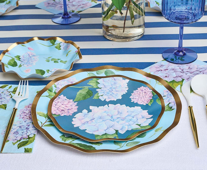A table set with a blue and white striped runner, Sophistiplate Salad Plate Hydrangea - 8pk, gold-edged cutlery, blue glassware, and a floral vase brings elegant spring charm to any celebration.