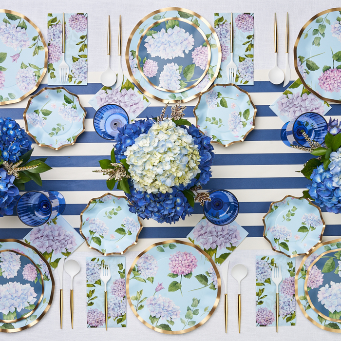 A table set with floral-patterned plates, gold cutlery, blue glasses, and Sophistiplate Cocktail Napkin Hydrangea 3 Ply atop a blue and white striped cloth, accented with blue and white hydrangea centerpieces.