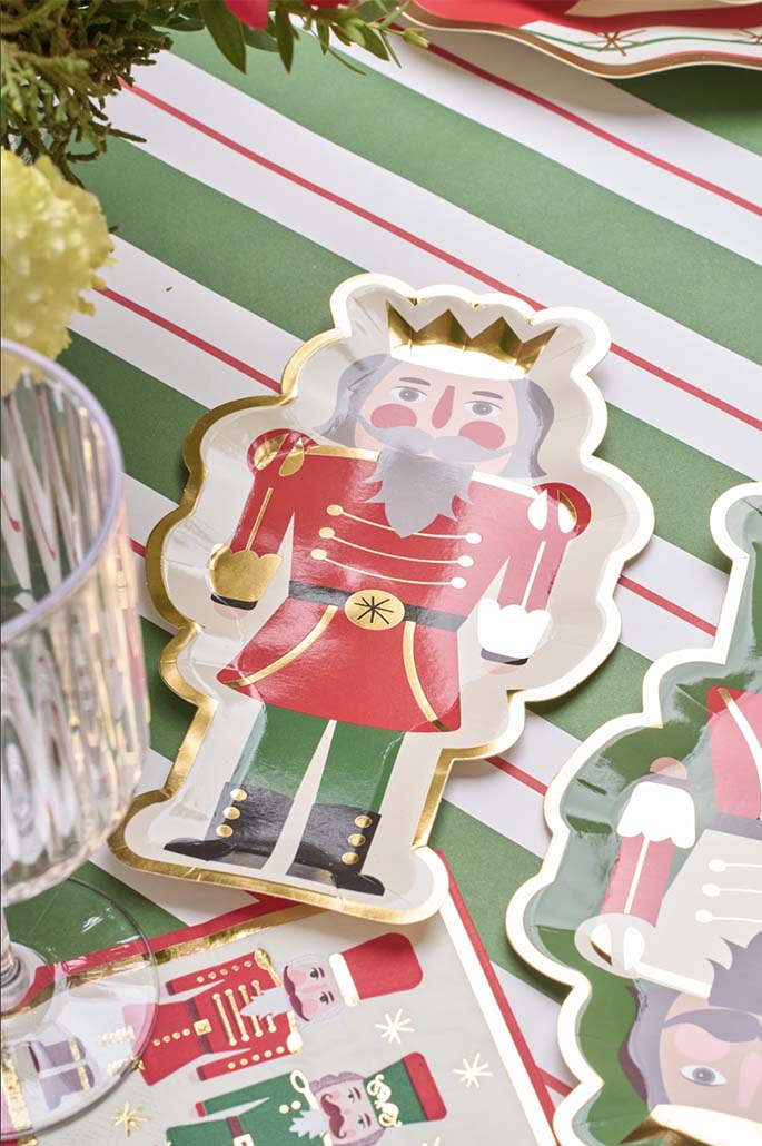 A festive table setting featuring the Sophistiplate Salad Plate Nutcracker Christmas Assorted (8/pkg) with red, gold, and green details on a green and white striped tablecloth. A clear glass and flowers are also partially visible.