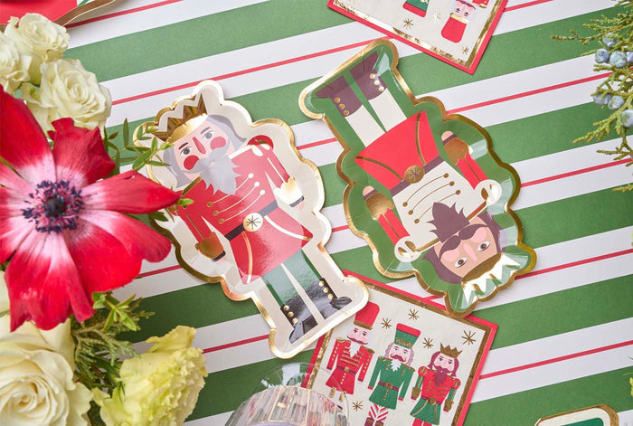 Create a festive holiday table with the Nutcracker Christmas Table Setting by Sophistiplate, featuring designer nutcracker-shaped disposable plates, nutcracker napkins, red and white flowers, and a green-and-white striped tablecloth.
