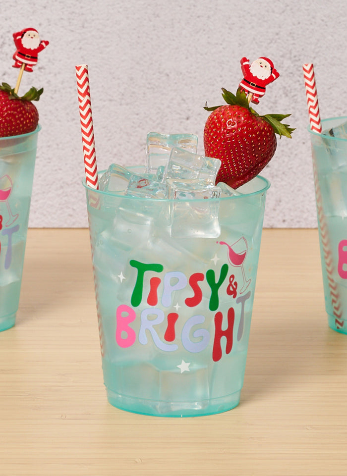 A turquoise cup with "Tipsy & Bright" text is filled with ice, a red and white striped straw, and a strawberry garnished using Simply Serv'd Santa Large Cocktail Picks (30pk). Two similar cups are partially visible in the background.