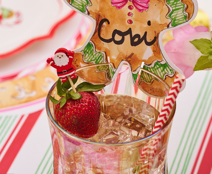 A glass with ice and a red-and-white striped straw holds a strawberry topped with a Simply Serv'd Santa Large Cocktail Pick. A gingerbread-shaped place card labeled "Cobi" sits on the rim, with festive tableware in the background.