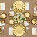 The Satin Gold Table Setting by Sophistiplate LLC features designer disposable plates, gold utensils, scalloped napkins, clear glasses, and a white and peach flower centerpiece on a brown textured tablecloth.