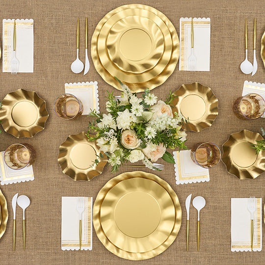 The Satin Gold Table Setting by Sophistiplate LLC features designer disposable plates, gold utensils, scalloped napkins, clear glasses, and a white and peach flower centerpiece on a brown textured tablecloth.