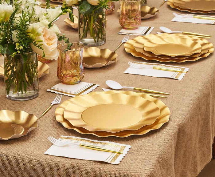 The Satin Gold Table Setting by Sophistiplate LLC features designer disposable plates, gold flatware, decorative glasses, white napkins with gold trim, and floral bouquets atop a burlap tablecloth for an elegant formal meal.