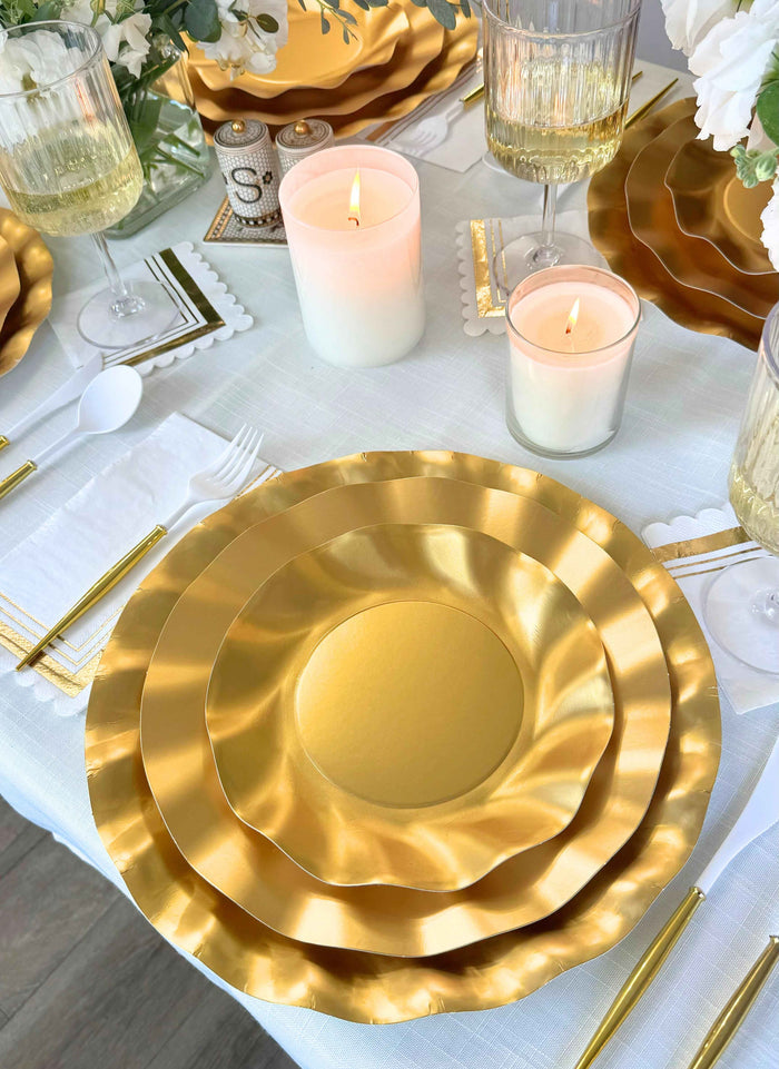 The Satin Gold Table Setting by Sophistiplate LLC includes designer disposable plates with gold wavy edges, gold flatware, crystal glasses, white napkins, and candles—perfect for an elegant party-in-a-box experience.