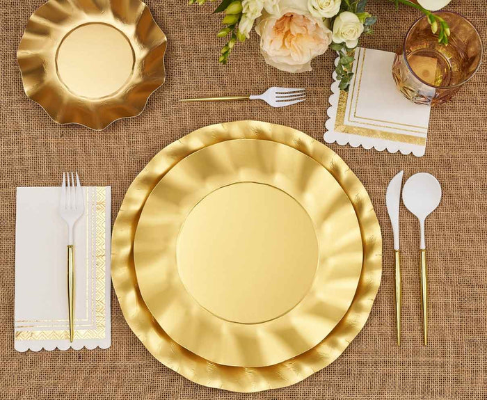 The Satin Gold Table Setting by Sophistiplate LLC features designer disposable plates, gold and white cutlery, a glass, elegant napkins with gold patterns, and a floral touch on burlap for a chic party-in-a-box look.
