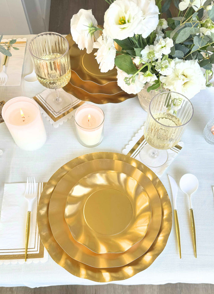 The Satin Gold Table Setting by Sophistiplate LLC includes designer plates, gold-rimmed glasses, gold-trimmed napkins, white candles, a floral centerpiece, and a white tablecloth—perfect for an elegant party-in-a-box.