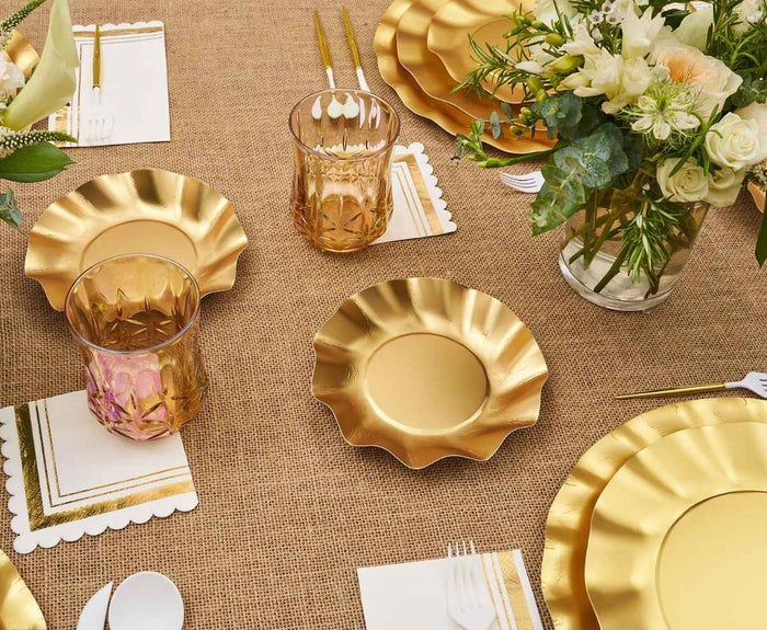 The Satin Gold Table Setting by Sophistiplate LLC features designer gold-rimmed disposable plates, clear geometric glasses, white napkins with gold edges, white plastic forks, and a bouquet of white flowers on a burlap tablecloth.
