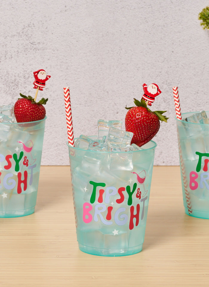 Three blue glasses with "Tipsy & Bright" text, filled with ice and garnished with a strawberry and Santa stir stick. Scarlet Chevron Paper Cocktail Straws by Simply Serv'd add a festive touch—makes a great hostess gift! Glasses sit on a light wood surface.