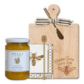Introducing the Shana Tova Serving Set by Sophistiplate LLC: a beautifully arranged Rosh Hashanah gift featuring a wooden board adorned with “Shana Tova” in English and Hebrew, a decorative bow, a honey jar with dipper, honeycomb-patterned napkins, and charming bee-themed accents.