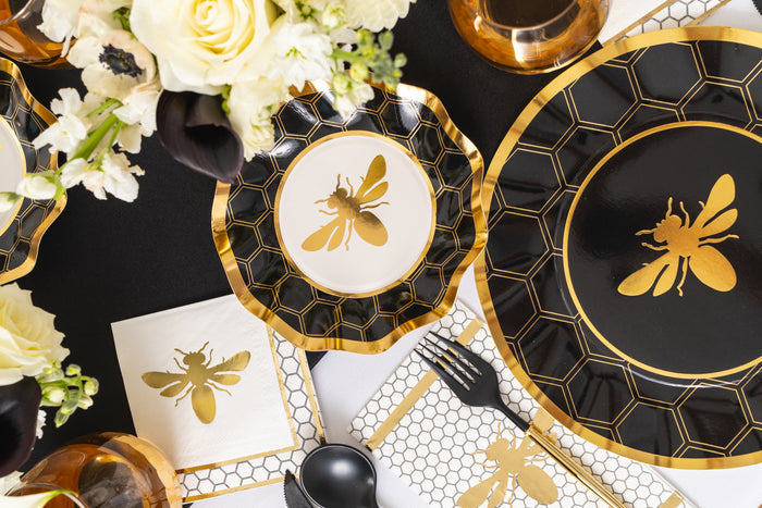A black and gold table setting features Sophistiplate Honeybee Dinner Plates in an 8-pack, highlighted by black and white party plates with ruffled edges, coordinating napkins, and decorations adorned with a charming honeybee design. The look is finished with honeycomb patterns, white flowers, and gold utensils.