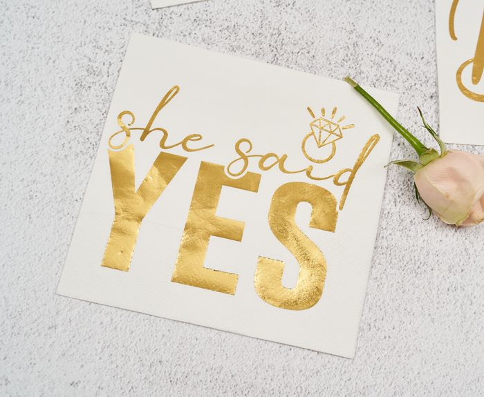 Sugarplum’s She said Yes Cocktail Napkins (26pk) feature shiny gold lettering and a gold diamond ring, perfect for bridal showers. Displayed beside a pale pink rose on a light textured surface.