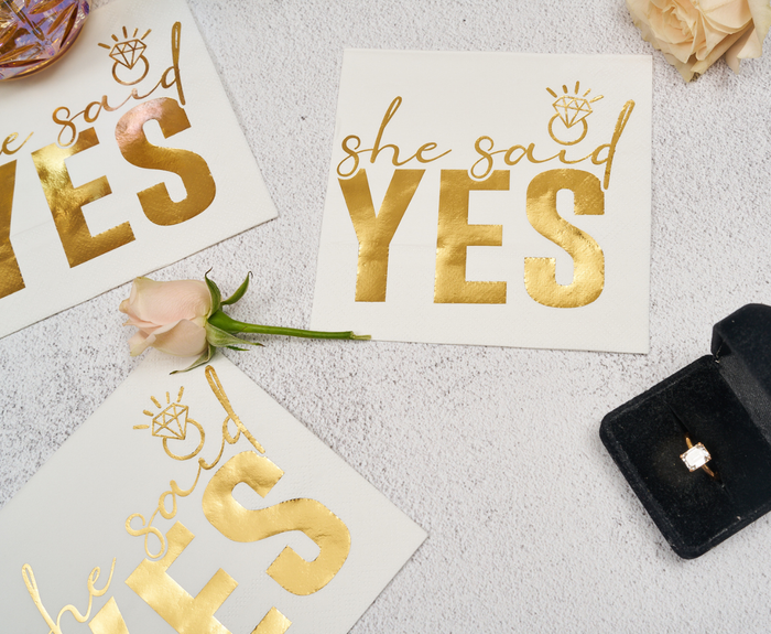 Sugarplum "She said Yes" Cocktail Napkins (26pk) feature gold text and diamond ring designs—ideal for bridal showers, bachelorette parties, or celebrations. Displayed with roses and a ring box for a stylish touch.