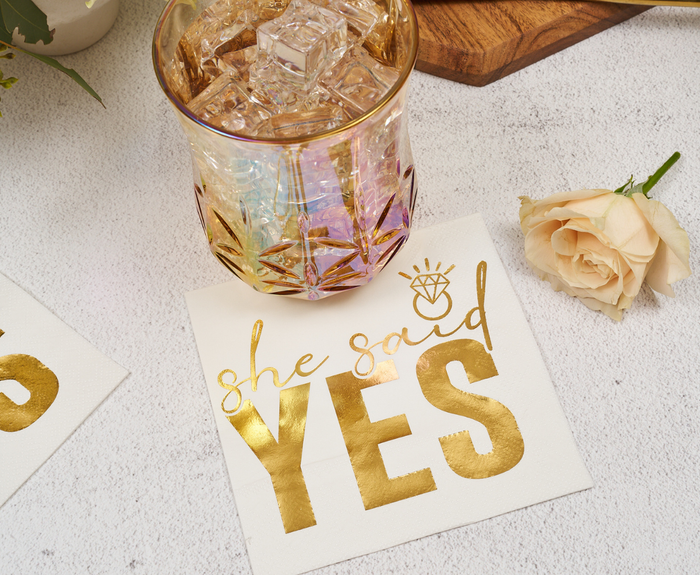 A glass with ice sits beside Sugarplum's "She said Yes" Cocktail Napkins (26pk), featuring bold lettering and a diamond ring graphic, with a cream rose nearby on a light surface.