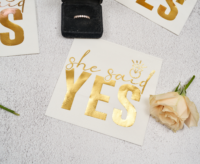Sugarplum’s "She said Yes" Cocktail Napkins (26pk) feature elegant gold lettering, shown here beside a blush rose and diamond ring box atop a light textured surface.