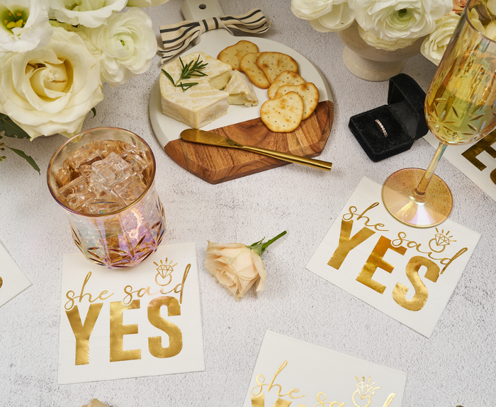 A festive scene features Sugarplum “She said Yes Cocktail Napkins” (26pk), champagne, whiskey on ice, cheese and crackers on a wooden board, a flower, a ring box, and white roses.