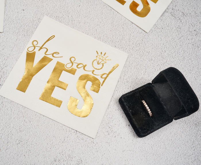 Sugarplum's She said Yes Cocktail Napkins (26pk) feature gold lettering and a diamond ring design—perfect bridal shower napkins, shown beside a black box with a gold diamond-studded ring on a textured light surface.