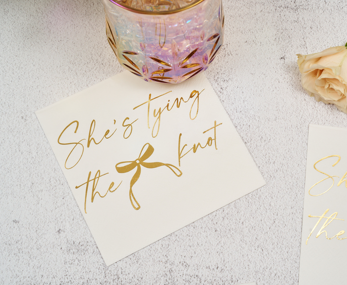 Sugarplum’s “She’s tying the Knot” Cocktail Napkins (26pk) are displayed next to a pink iridescent glass and a pale peach rose on a light textured surface.