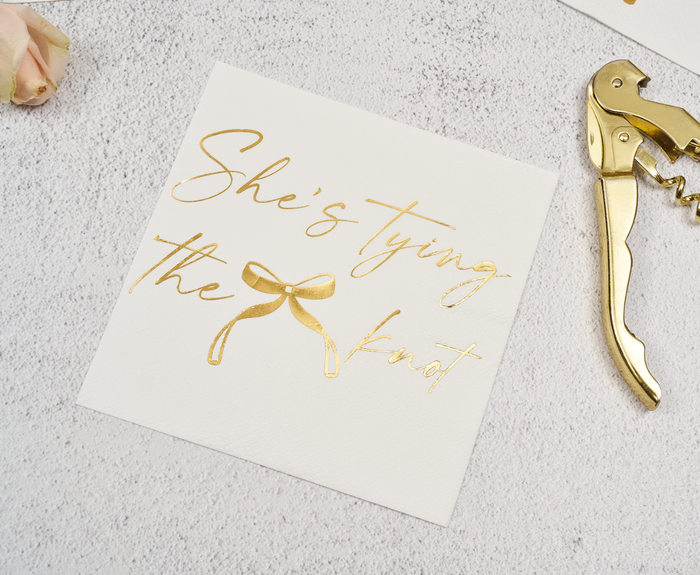 Sugarplum's "She's tying the Knot" Cocktail Napkins (26pk) feature gold foil script and a bow design—perfect bridal shower or bachelorette party napkins. Shown on a textured surface beside a gold corkscrew and pale pink rose.