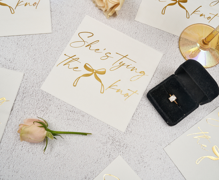 A flat lay featuring Sugarplum’s “She’s tying the Knot” Cocktail Napkins (26pk), blush roses, a champagne glass, and an open ring box displaying a diamond engagement ring.