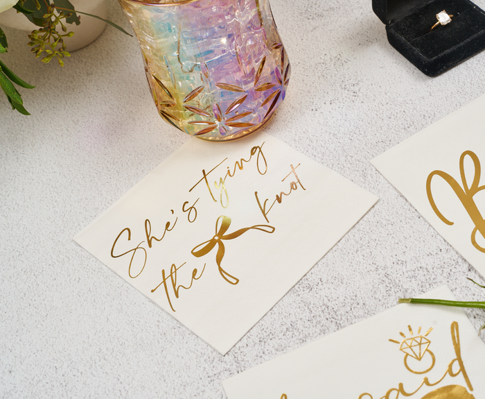 Sugarplum’s “She’s tying the Knot” Cocktail Napkins (26pk) are displayed with a ring box, pastel iridescent glass, floral accents, and gold script card on a light surface for an elegant bridal setup.