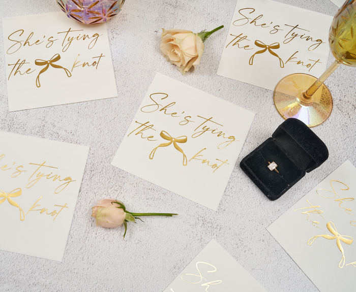 Flat lay of Sugarplum She's tying the Knot Cocktail Napkins (26pk), gold-rimmed glasses, cream roses, and cards with gold script around a black ring box with an engagement ring on a light textured background.