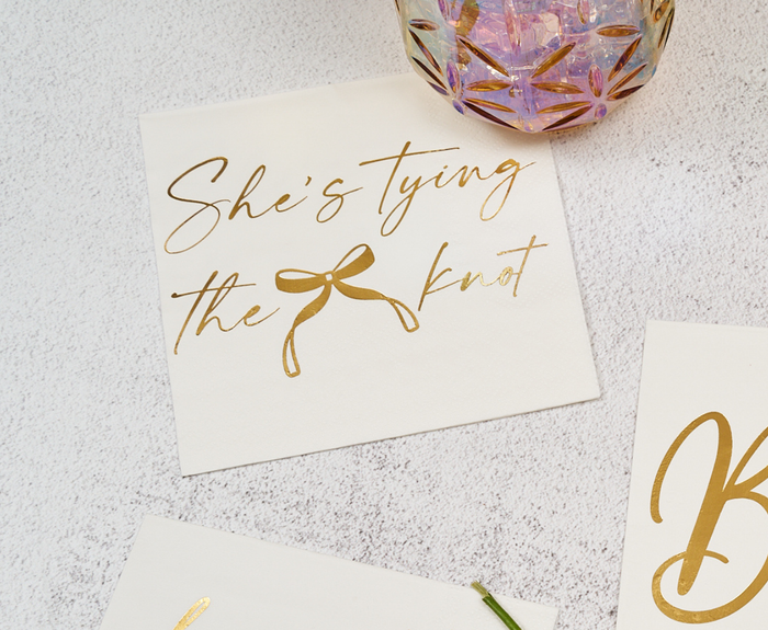 The Sugarplum "She's tying the Knot" Cocktail Napkins (26pk) feature elegant script and are ideal for bridal showers, pictured with a patterned glass and another napkin on a light surface.