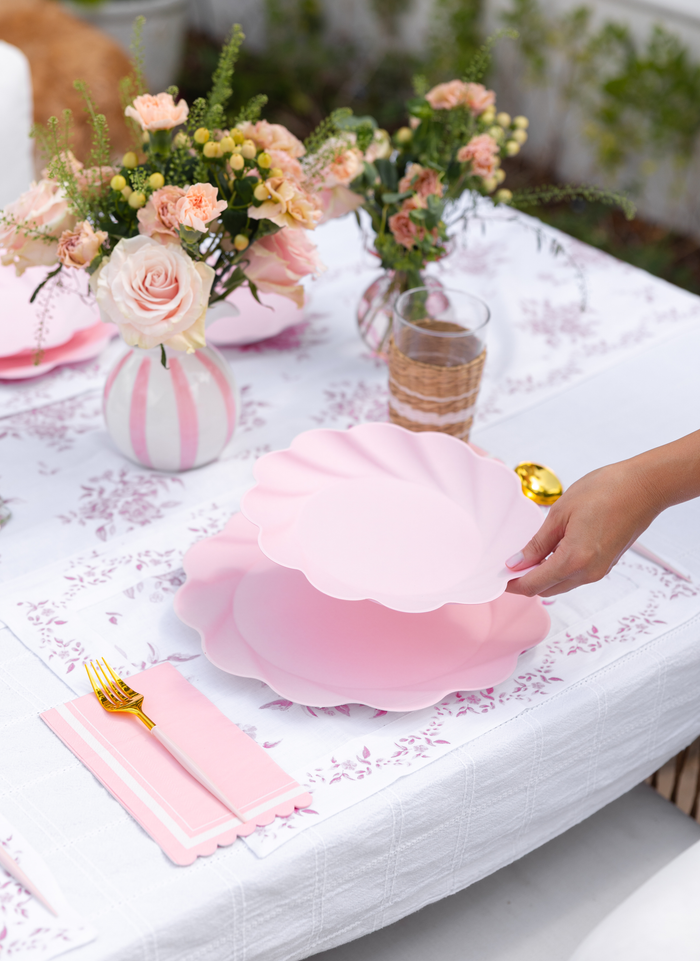 A hand sets Sophistiplate LLC's Eco-Friendly Table Setting- Blush—pink, scalloped disposable plates—on a white table with floral decor, gold cutlery, and pink napkins for an elegant, festive look. A wicker glass sits beside the stylish arrangement.