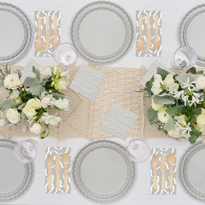 A top-down view of a table set for four with eco-friendly Simply Eco Dinner Plate-Concrete by Sophistiplate, gold cutlery on patterned napkins, striped paper napkins, white floral bouquets, purple glasses, and a cream woven runner.