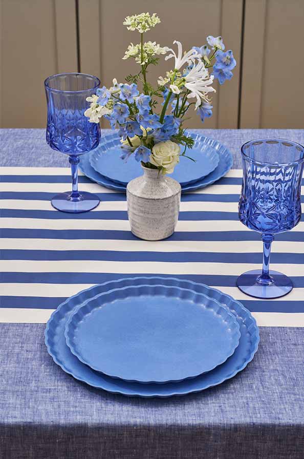 The Sophistiplate Eco-Friendly Table Setting- French Blue features blue scalloped plates, two blue patterned wine glasses, and a small vase of white and blue flowers on a blue tablecloth with a white and blue striped runner.