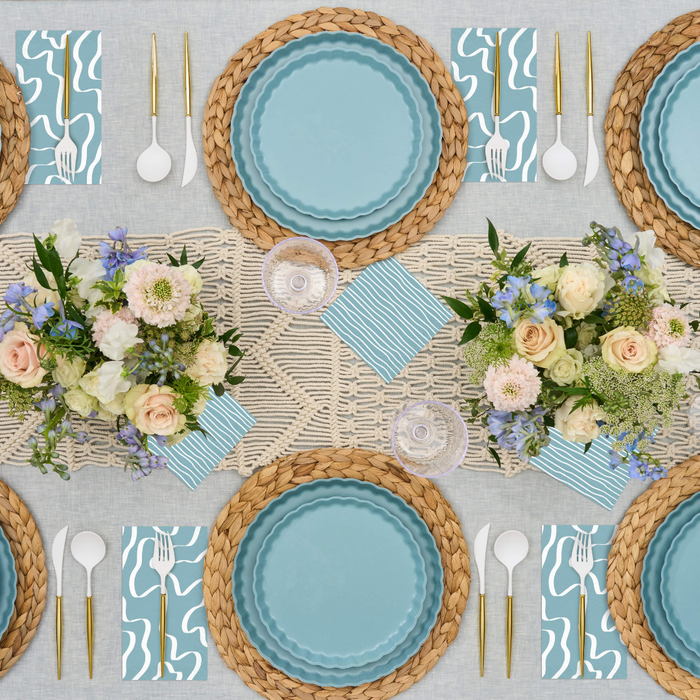 A top-down view of a table set for four with Sophistiplate's Simply Eco Dinner Plate- Pool, woven chargers, gold and white cutlery, blue-patterned napkins, floral centerpieces, and a macramé runner on a neutral tablecloth.