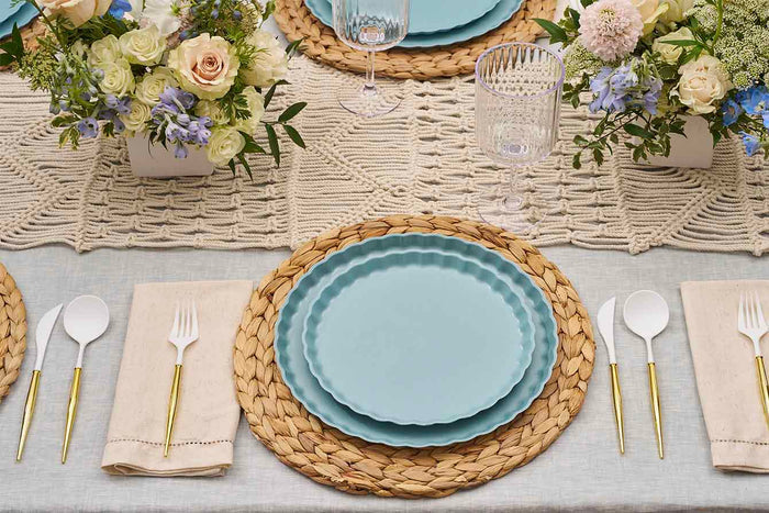 A table setting with a woven placemat, Sophistiplate Simply Eco Dinner Plate- Pool, gold-handled cutlery, cream napkins, and floral centerpieces on a light tablecloth with a lace runner.