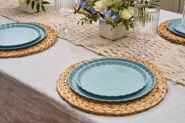 A close-up of a meal table set with Sophistiplate Simply Eco Dinner Plate-Pool, woven placemats, clear ribbed glasses, a crochet runner, and white and blue floral centerpieces.