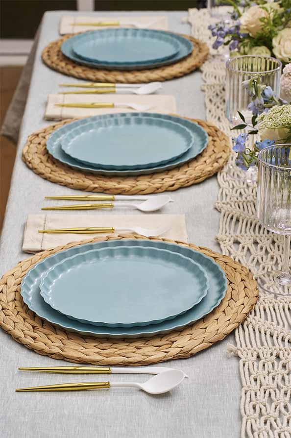 A neatly set dining table with woven placemats, Sophistiplate Simply Eco Salad Plates in Pool, gold-handled cutlery, white napkins, clear ribbed glasses, and a macramé table runner decorated with floral arrangements.
