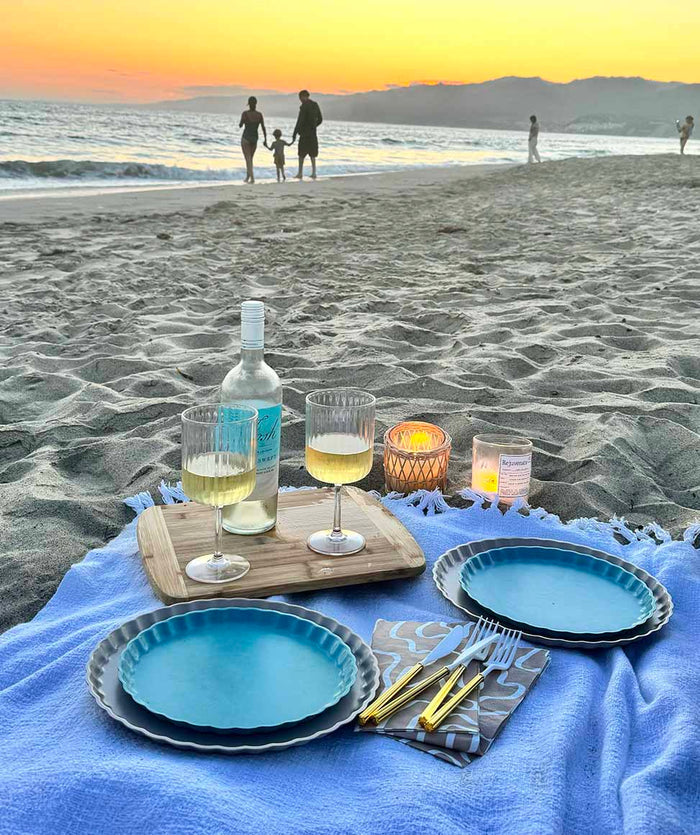 At sunset, a Sophistiplate Eco-Friendly Table Setting- Pool Blue is arranged on the sand with plates, wine glasses, a bottle of white wine, and cutlery. In the background, a family strolls along the shoreline by the ocean.