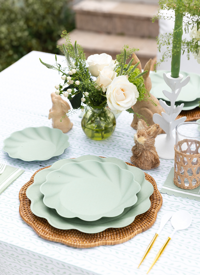 A spring-inspired outdoor table features Sophistiplate LLC’s Eco-friendly Table Setting- Sage with green disposable plates, white cutlery, a woven charger, white roses in a vase, a straw bunny figurine, and a green candle on display.