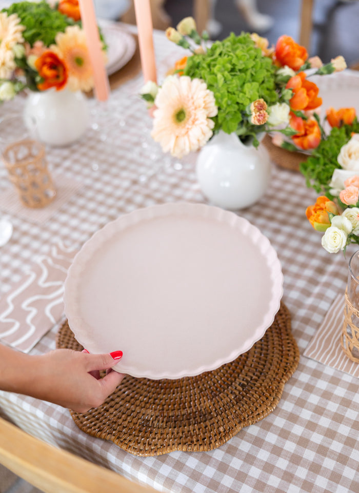 A hand sets a Simply Eco Dinner Plate- Sand/8pkg by Sophistiplate, a round, pale pink compostable plate, on a woven placemat atop a checkered tablecloth amid floral arrangements, candles, and glasses for an eco-friendly gathering.
