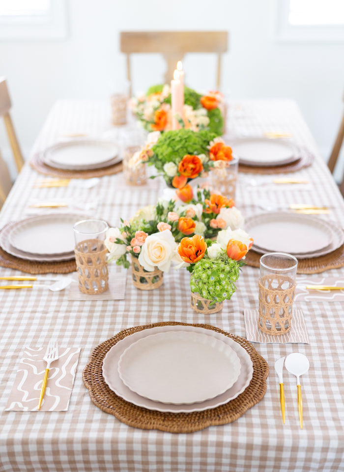 A dining table set with Sophistiplate’s Simply Eco Dinner Plate in Sand, beige plates on woven placemats, gold utensils, and glasses with wicker holders. A checkered tablecloth is adorned with orange and white floral centerpieces.