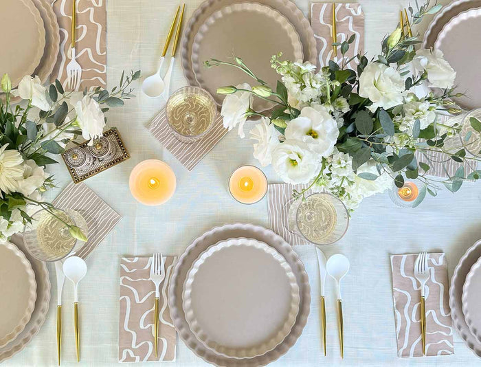 Elevate your table with Sophistiplate Simply Eco Dinner Plate- Sand (8/pkg), paired with gold cutlery, white plastic utensils, patterned napkins, white floral centerpieces, and candles on a cream tablecloth for an eco-friendly yet elegant setting.