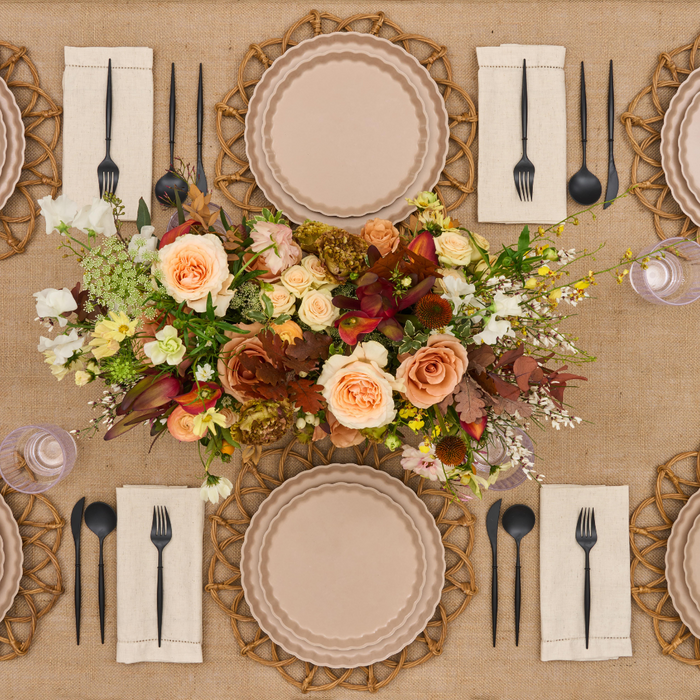 A top-down view of a table for six set with Sophistiplate Simply Eco Dinner Plate- Sand, black cutlery on cream napkins, water glasses, and a floral centerpiece with orange, yellow, and white blooms atop a burlap tablecloth.