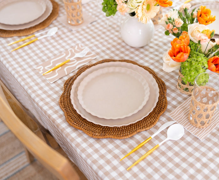 A table is set with Sophistiplate Simply Eco Dinner Plates in Sand, woven placemats, gold and white cutlery, clear glasses, and floral centerpieces of orange and cream blooms atop a checkered tablecloth.